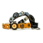 Arnés de cuero negro con parches de cuero amarillo y plateado, studs de latón y plata, ornamentos metálicos y un emblema decorativo, visto desde la izquierda.