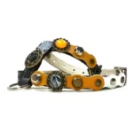 Arnés negro con parches amarillos y plateados, studs de metal mixto, detalles ornamentales y un emblema de esmalte negro, visto desde la derecha.